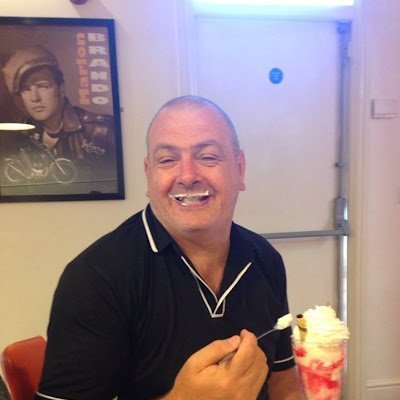 A man sitting at a table with a dessert, smiling and holding a spoon, has whipped cream on his upper lip. A vintage poster of a man in the background.